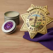 A 5-gram TrueBadge Wax tin with purple Owl Badges label sitting open next to a gold-plated San Mateo County Deputy Sheriff's Office star badge, displayed on a purple microfiber polishing cloth against a light wood background.